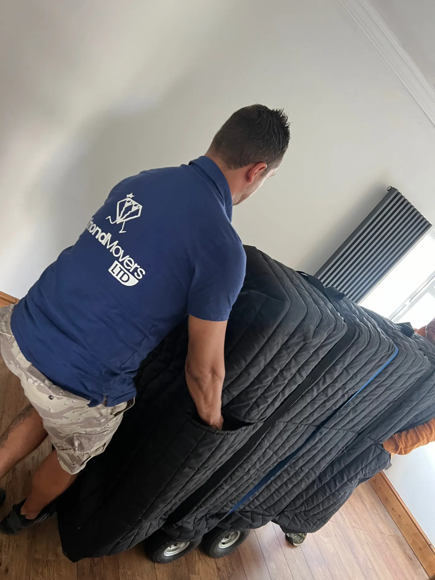 Dimond Movers mover in branded shirt manoeuvring a wrapped piano indoors
